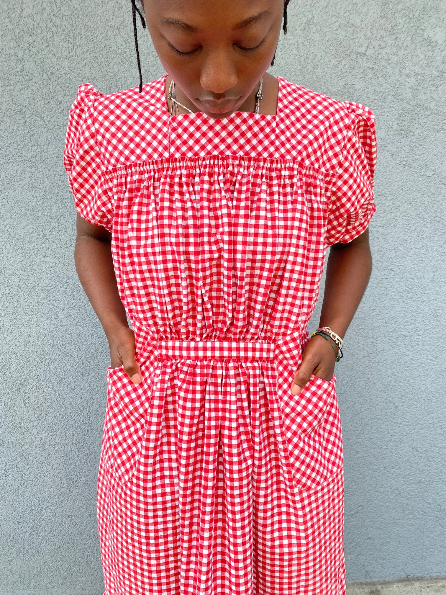 Close up of woman standing in front of grey wall wearing a red and white check dress with short puffed sleeves and a bottom flounce.