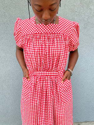 Close up of woman standing in front of grey wall wearing a red and white check dress with short puffed sleeves and a bottom flounce.