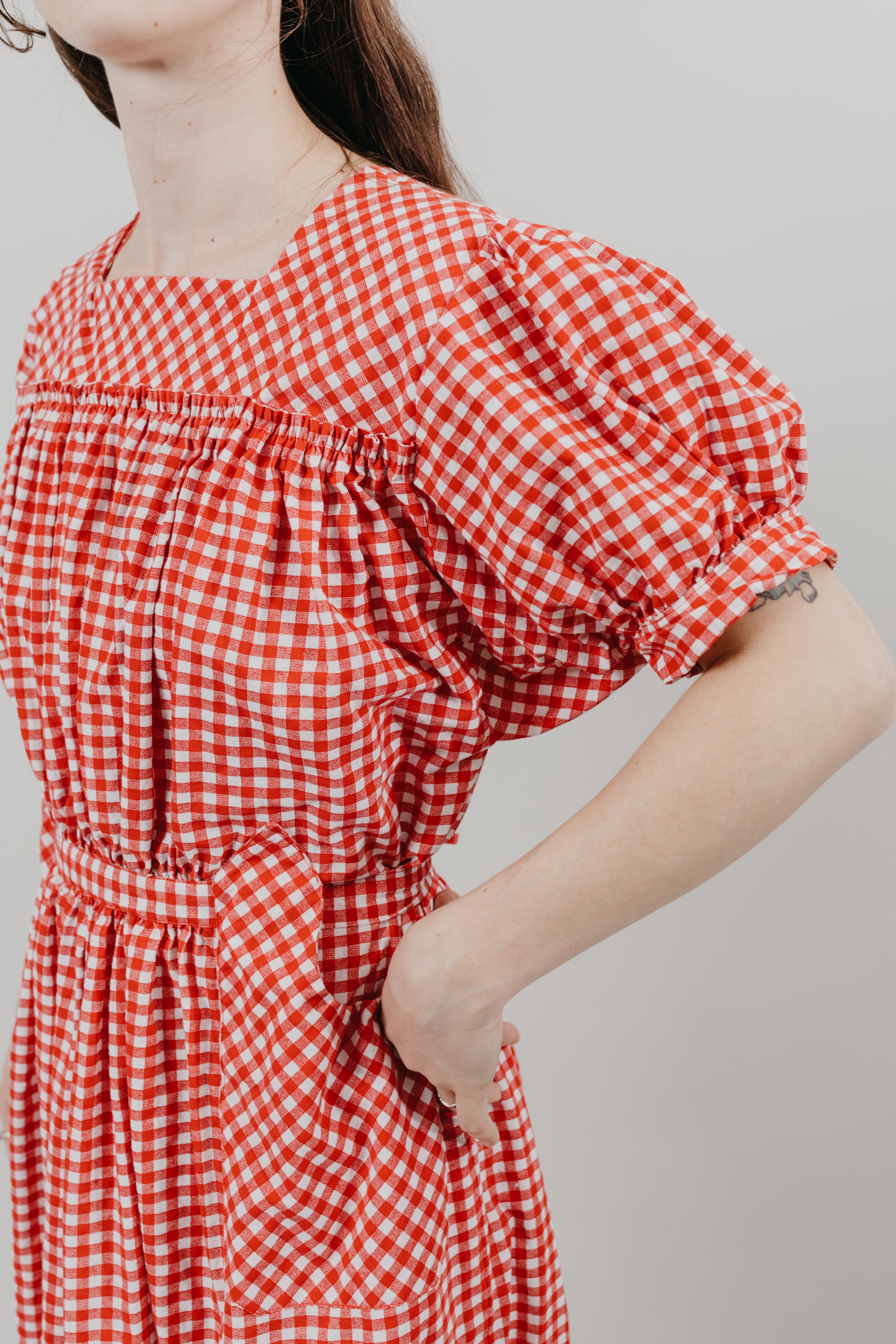 Close up of Japanese American woman standing in front of a white background wearing a red and white gingham calf-length dress with puffed sleeves, and a flounce.  Dress is belted at the waist and has pockets cut on the bias.