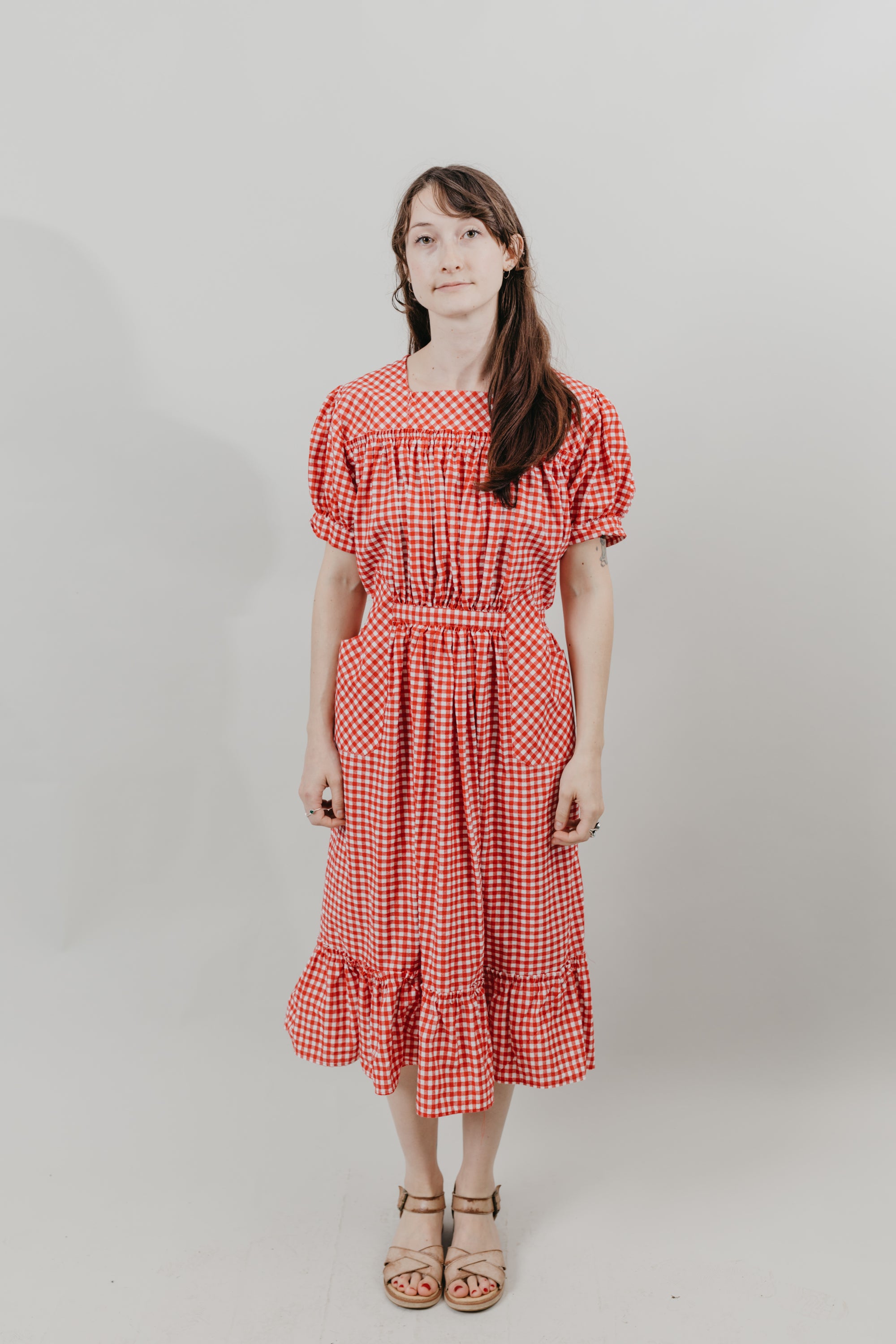 Japanese American woman standing in front of a white background wearing a red and white gingham calf-length dress with puffed sleeves, and a flounce.  Dress is belted at the waist and has pockets cut on the bias.