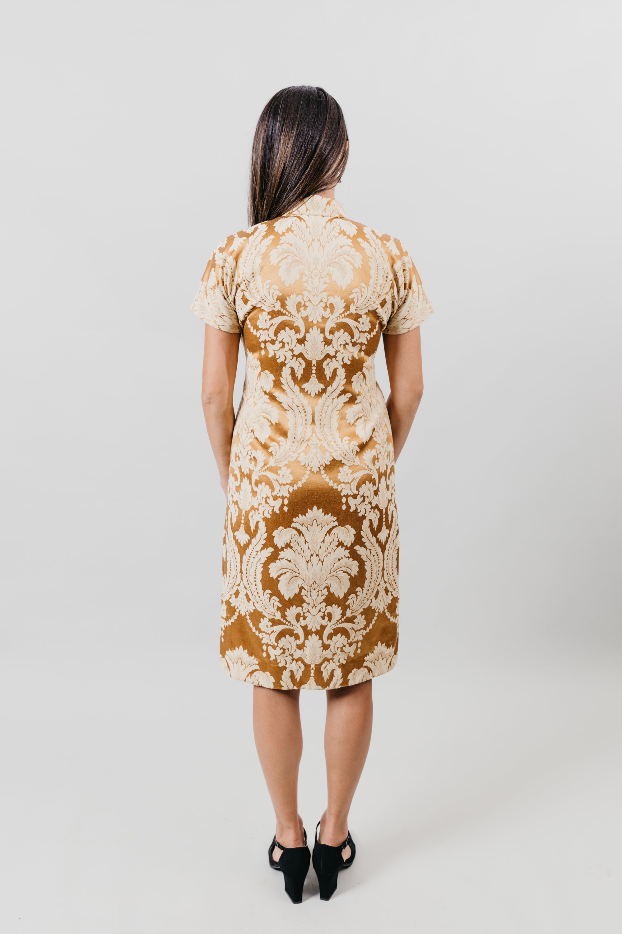 Back view of woman wearing knee-length gold brocade cheongsam in front of white backdrop