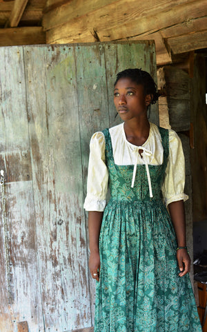 Young woman wearing green Dirndel (View B dress with View A Blouse). She is standing in front of a wood door. Model is looking over her right shoulder with her hands by her sides.