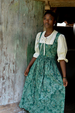 Young woman wearing green Dirndl (View B dress with View A Blouse).  She is standing in front of a wood door holding skirt in both hands by her side.