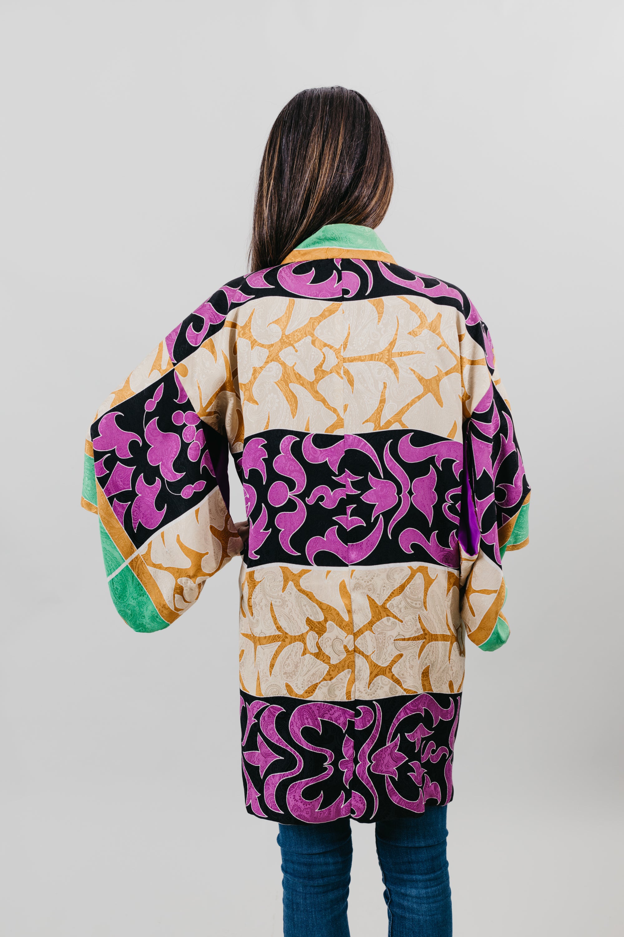 Asian woman wearing a purple, black, green, and gold printed Haori - back view with white back ground.