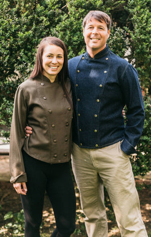 Man and woman standing together wearing a navy and brown Belgian Military Chef's Coats, respectively.  Outside by bushes.