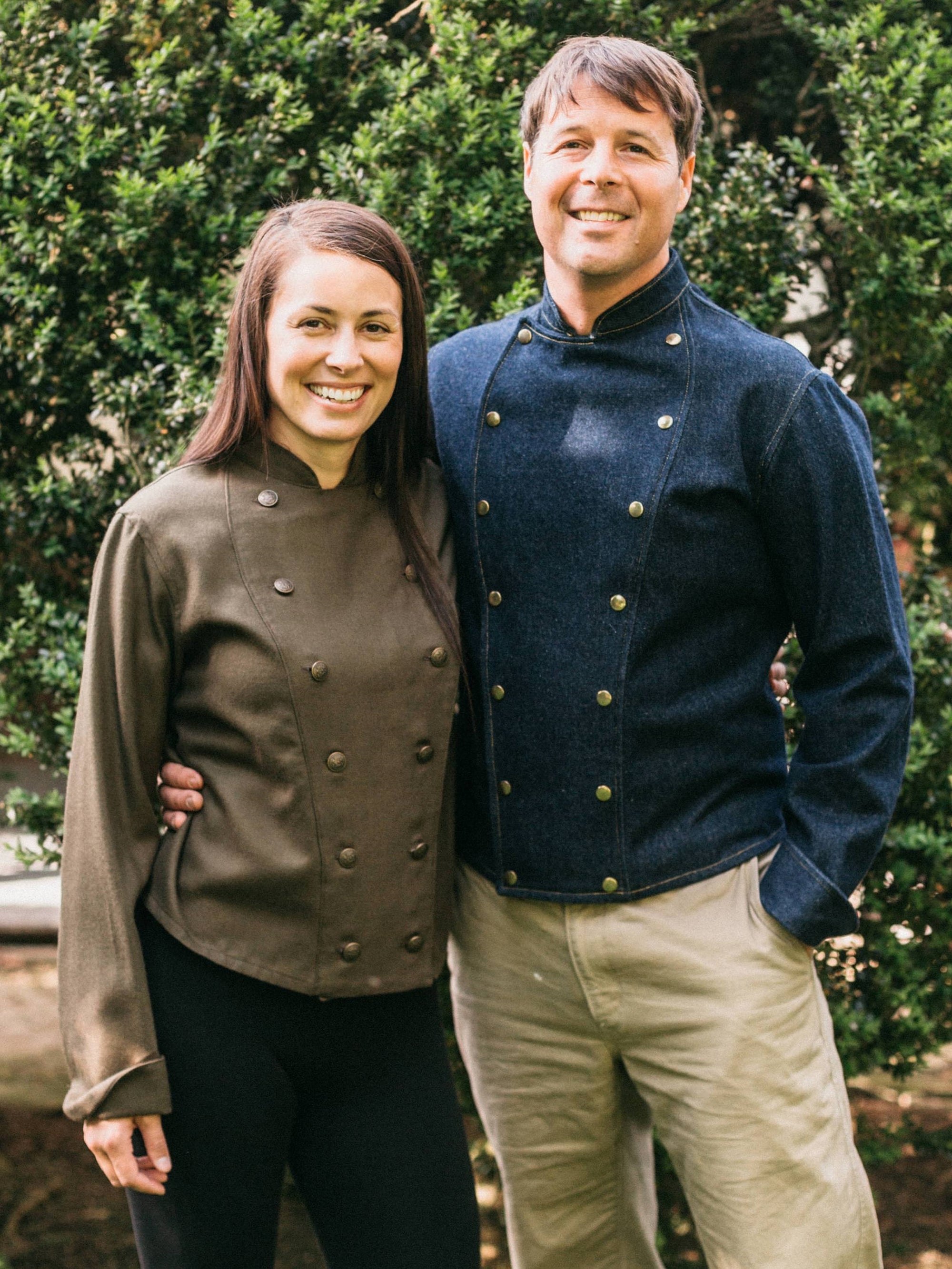 Man and woman standing together wearing a navy and brown Belgian Military Chef's Coats, respectively. Outside by bushes.