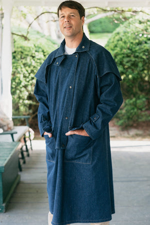 Man standing on a porch wearing a dark blue Drover's Coat.  hands in pockets.