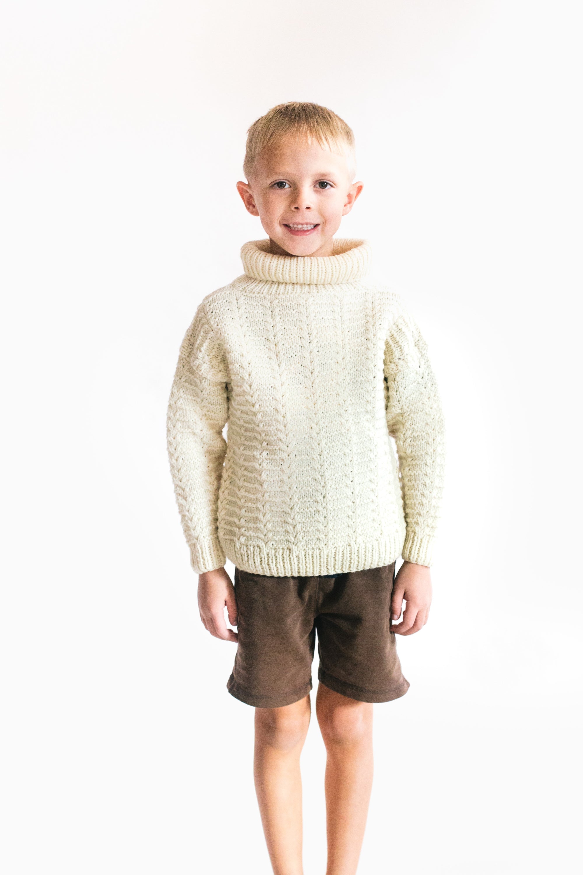 Small boy wearing a cream colored knitted sweater standing in front of a white screen