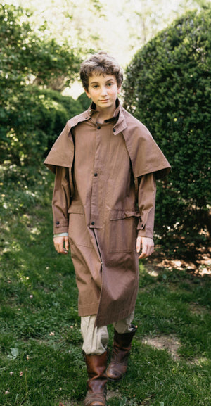 Older boy walking toward camera wearing a brown canvas Drover's Coat.  Outside in front of green bushes.