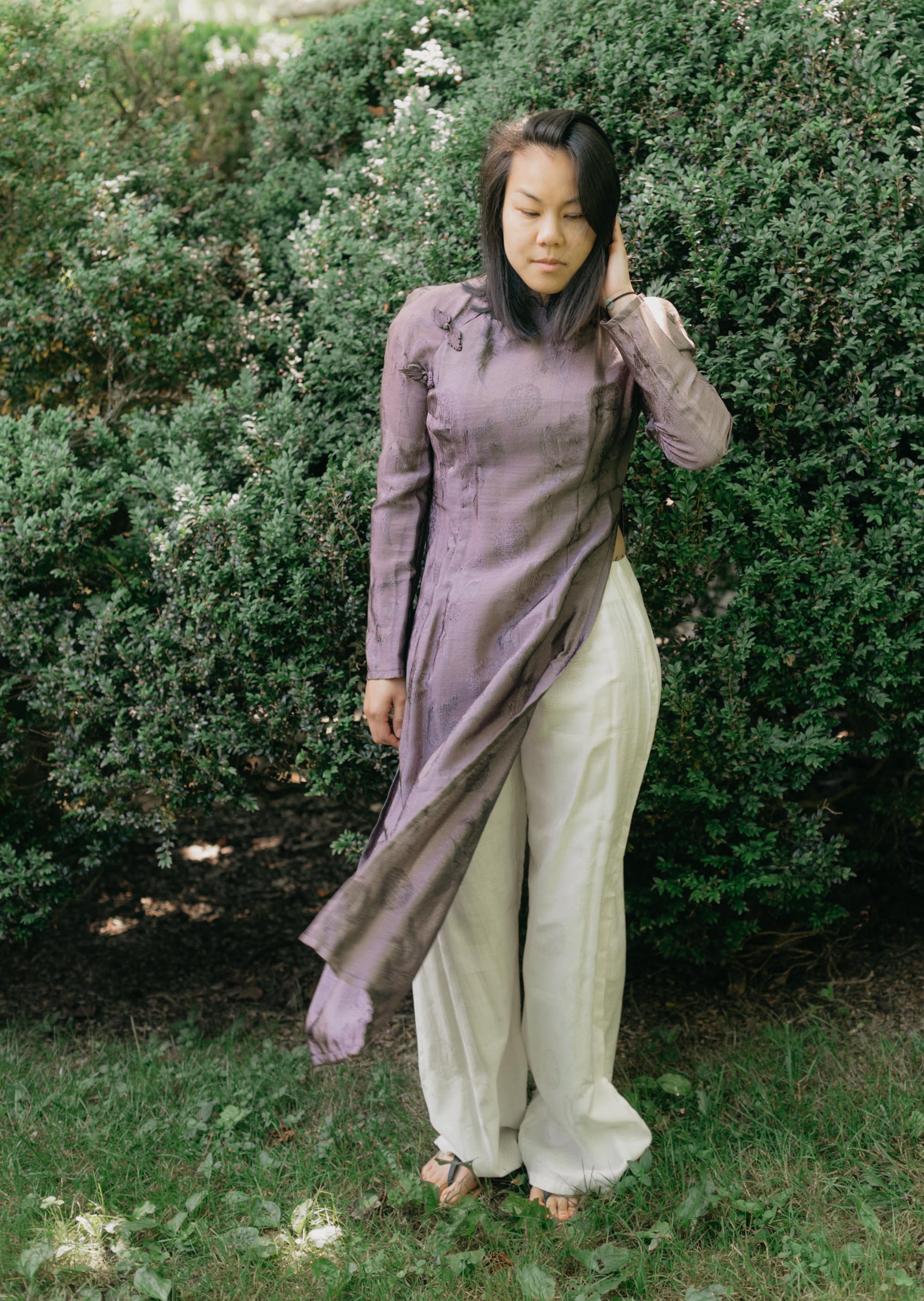 Vietnamese American woman wearing a purple silk Ao Dai tunic with white pants and sandals. She is standing in a lawn with shrubs in the background and her hand is at her hair.
