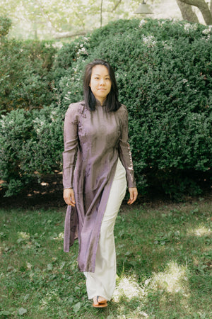 Vietnamese American woman wearing a purple silk Ao Dai tunic with white pants and sandals.  She is walking toward the camera in a lawn with shrubs in the background.