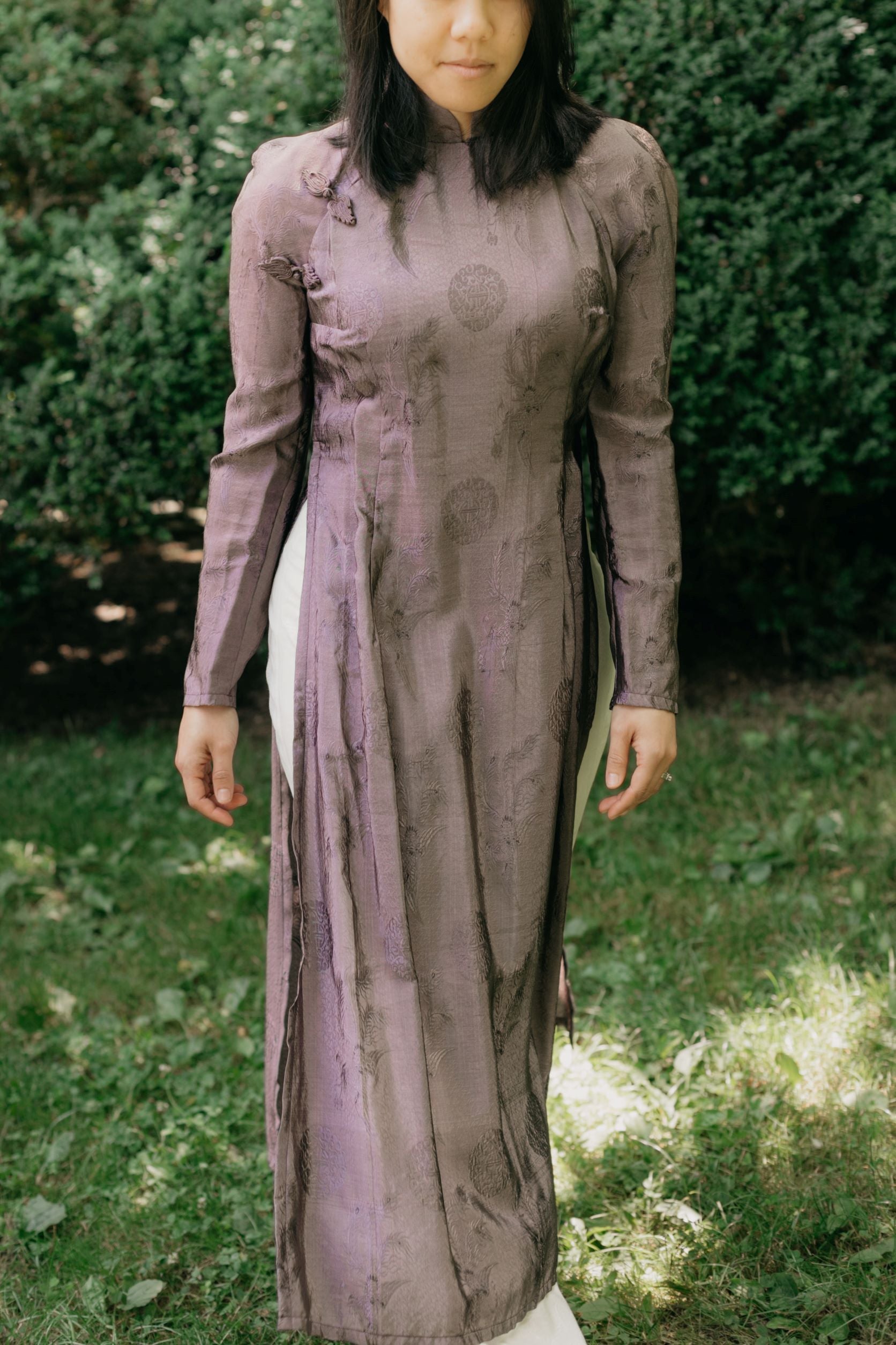 Vietnamese American woman wearing a purple silk Ao Dai tunic with white pants and sandals. She is walking toward the camera in a lawn with shrubs in the background.