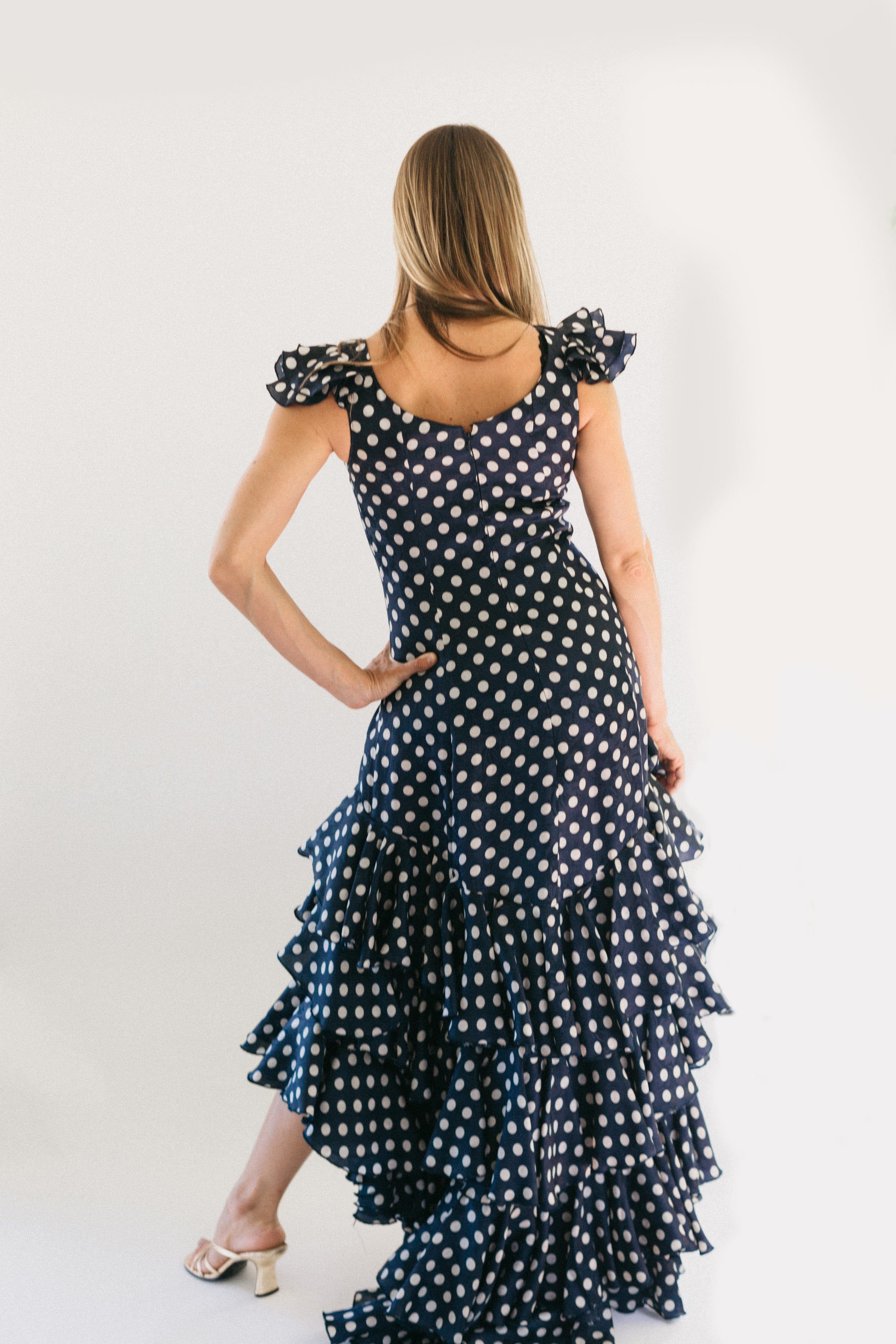 Photo of back view of woman wearing the Flamenco dress.  She stands in front of a white studio backdrop and has her left hand on her hip.