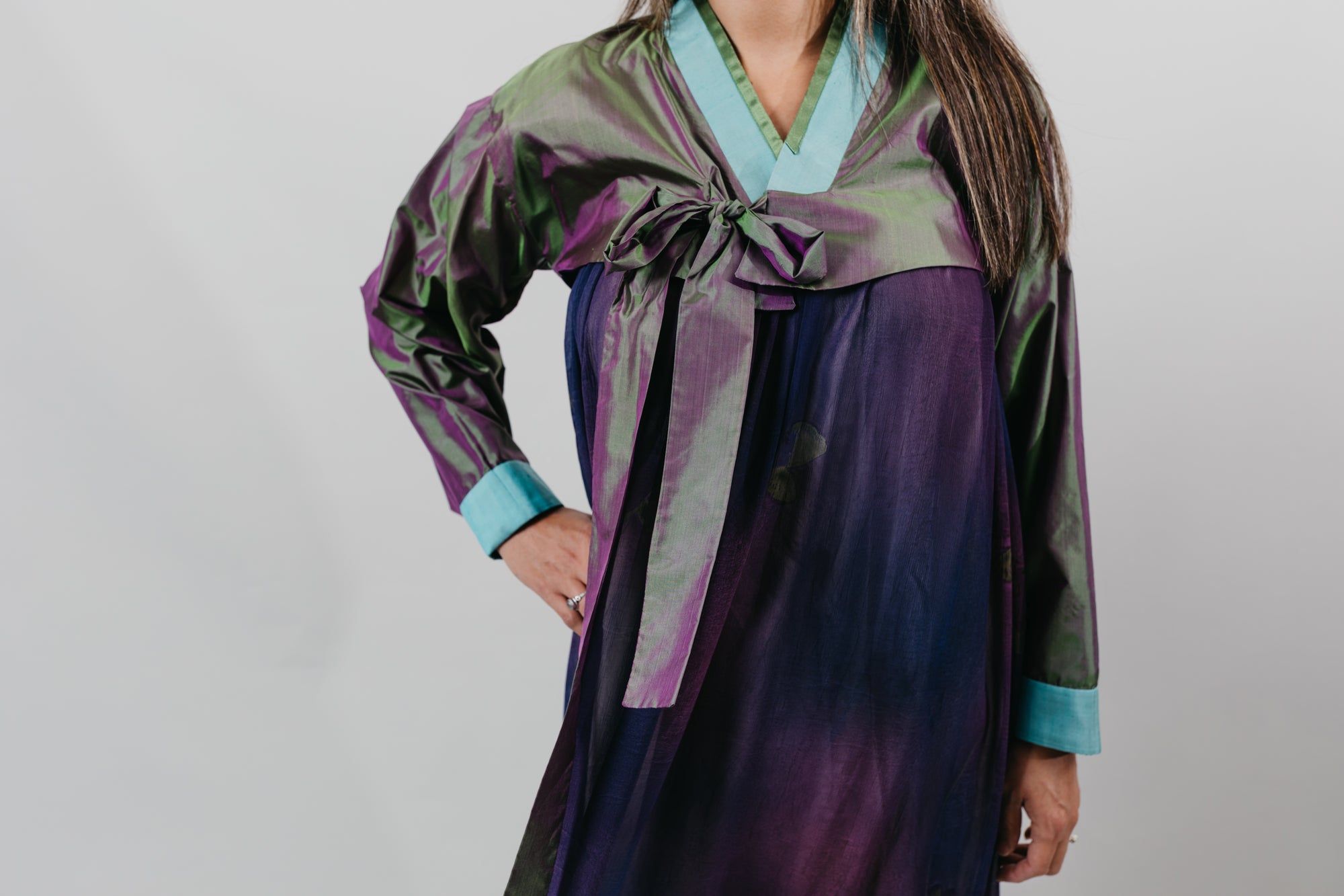 Woman wearing a purple shot silk han bok dress and jacket. Jacket is of teal and green shot silk and tied in the front. She is standing in front of a white backdrop.  Photo is a close up of the front of the hanbok