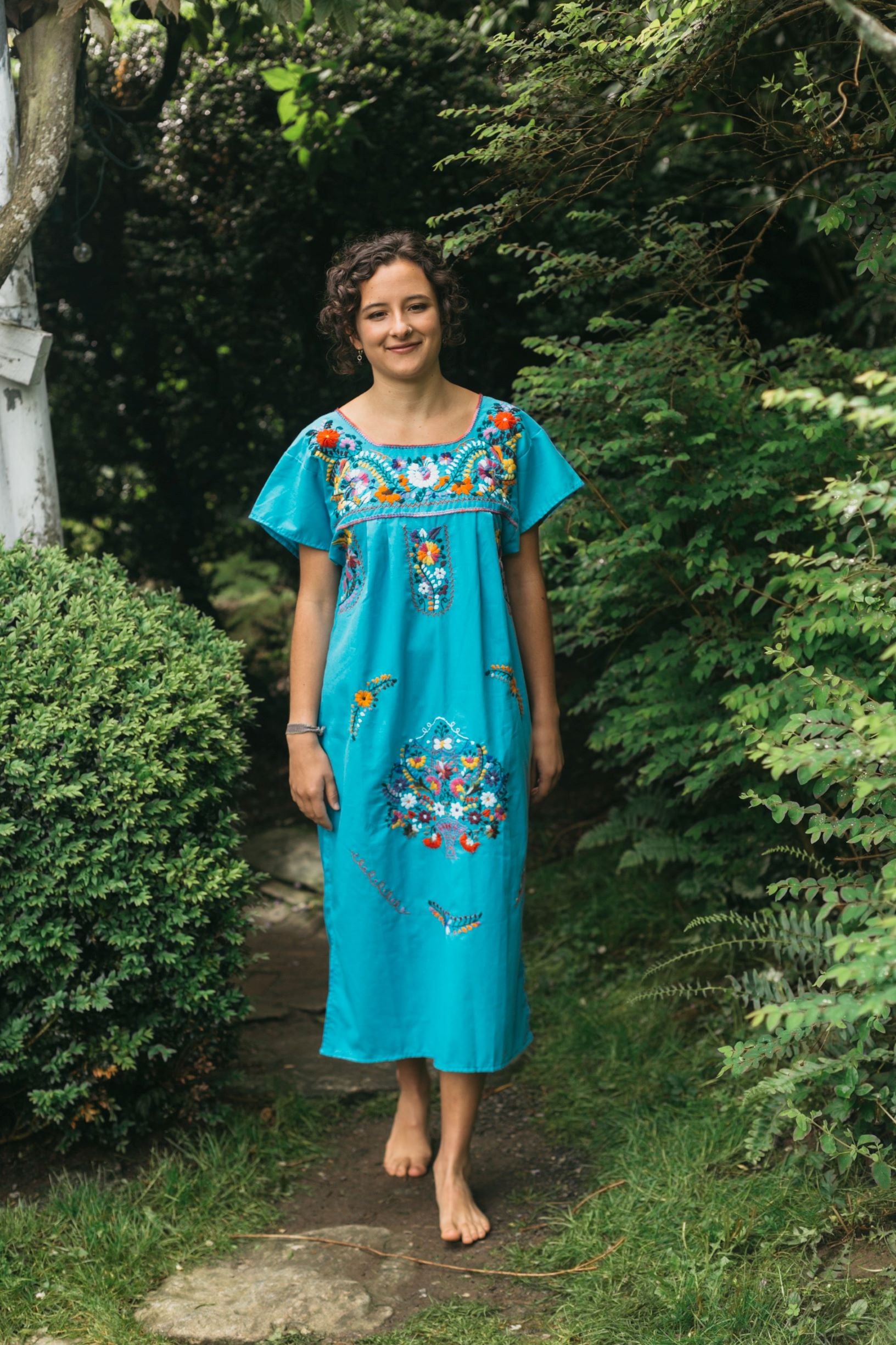 Photo of a young woman walking barefoot outdoors wearing a blue Old Mexico dress with colorful floral embroidery.