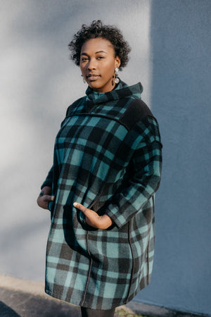 Black woman standing outside in front of a grey wall wearing a green and black plaid siberian parka.  Her hands are in the pockets.