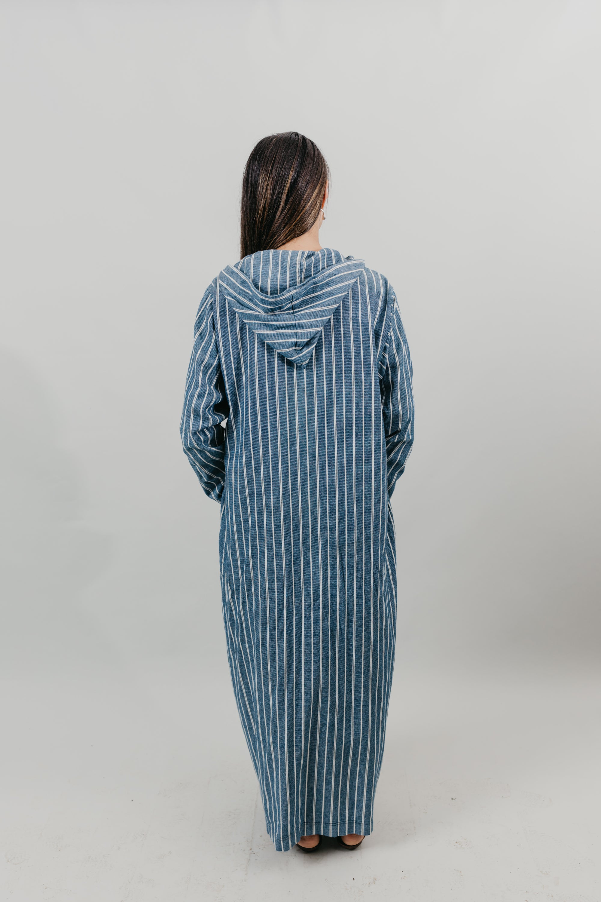 Asian woman wearing a floor length blue and white vertical striped Djellaba with her hands in her pockets.  Back view.  Background is white back drop.