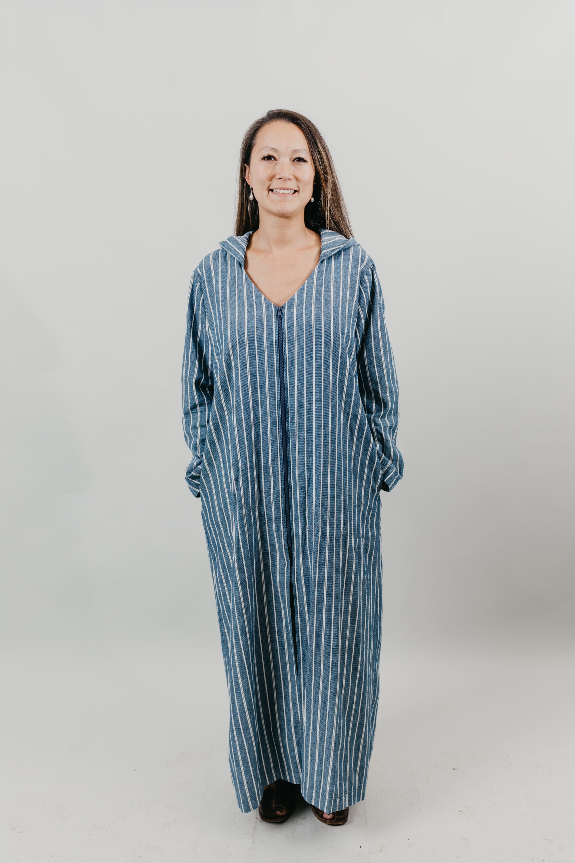 Asian woman wearing a floor length blue and white vertical striped Djellaba with her hands in her pockets.  Background is white back drop.