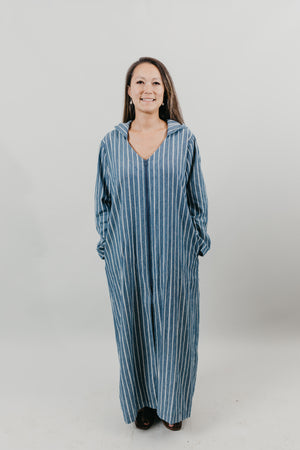 Asian woman wearing a floor length blue and white vertical striped Djellaba with her hands in her pockets.  Background is white back drop.