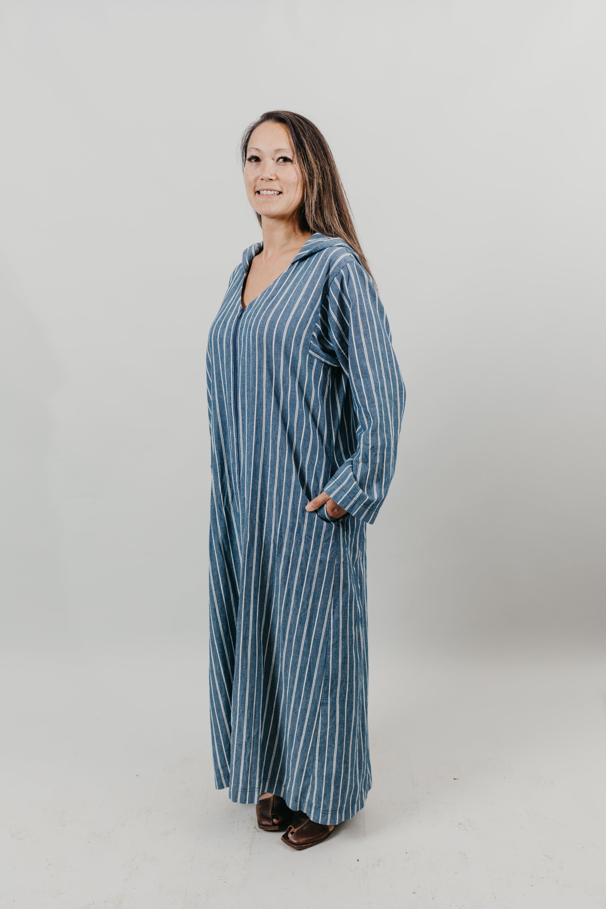 Asian woman wearing a floor length blue and white vertical striped Djellaba with her hands in her pockets.  She is turned slightly to the side.Background is white back drop.