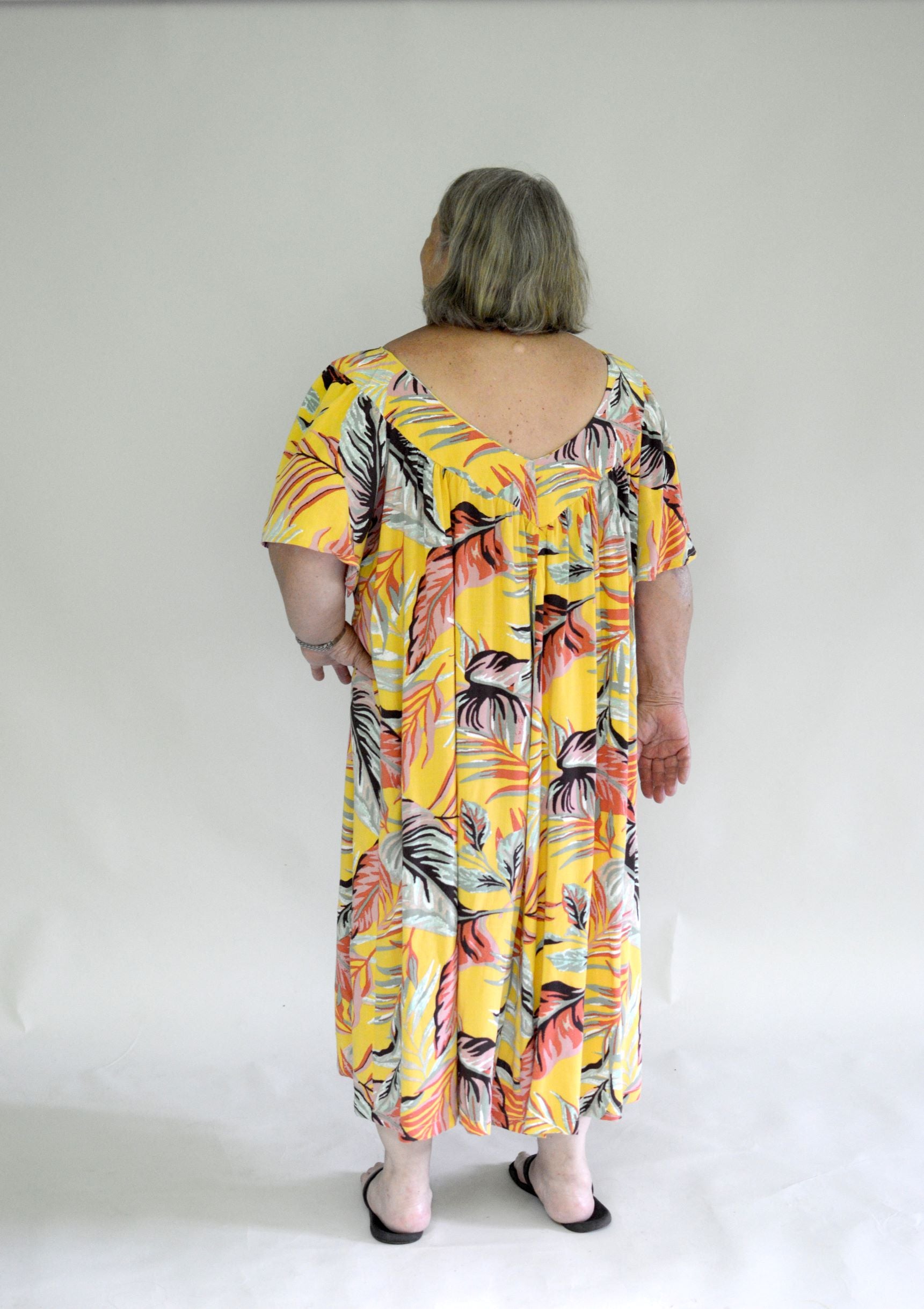 Back view of Older woman wearing a size 2XL knee-length, short-sleeve, yellow muumuu with orange and green palm fronds on it.