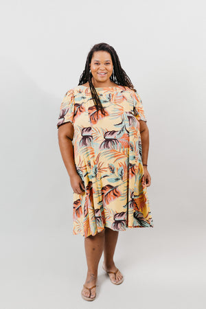 Black woman with long thin dreds, wearing a size 2XL knee-length, short-sleeve, yellow muumuu with orange and green palm fronds on it.  Background is white back drop.