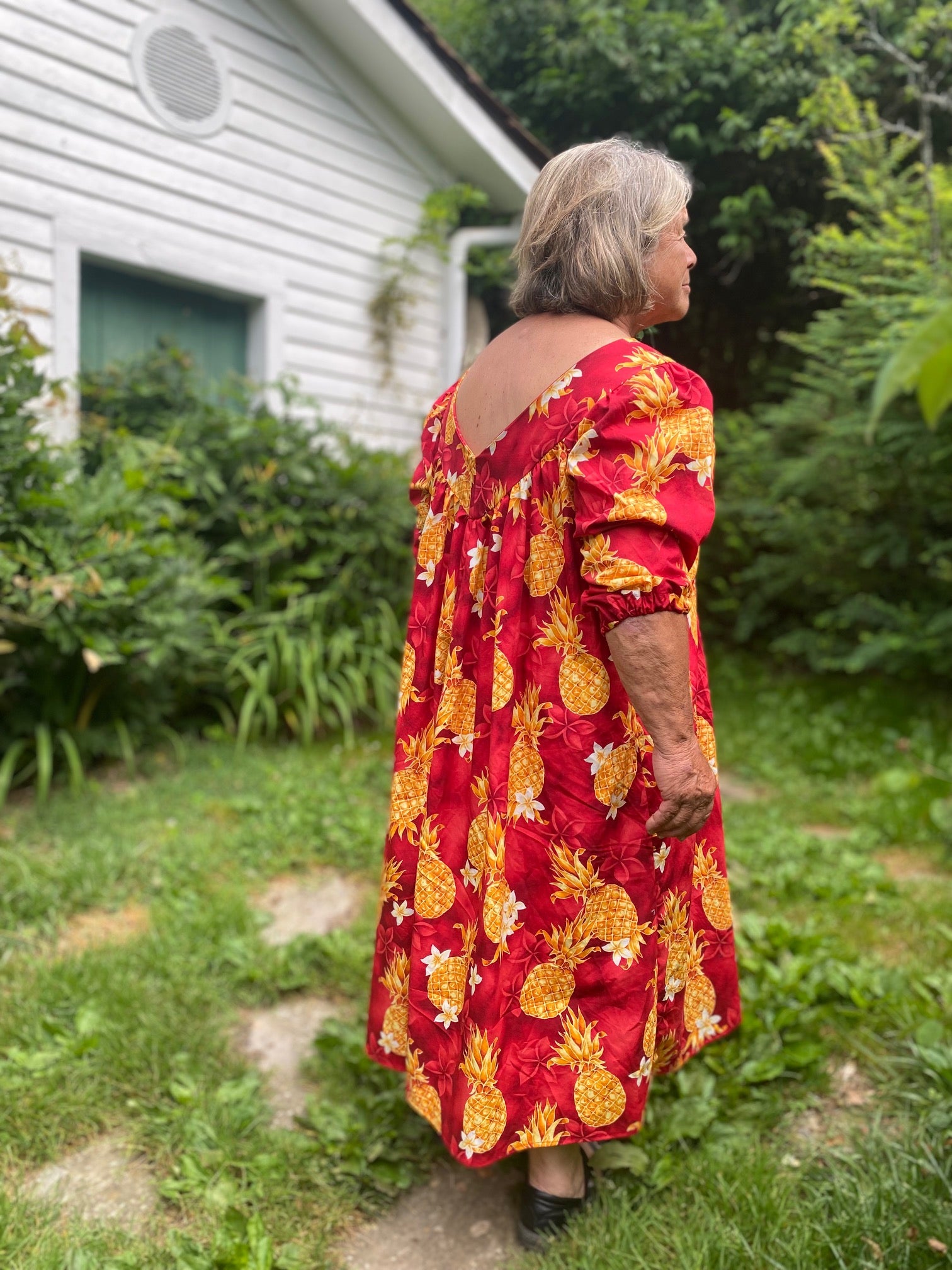 Older woman wearing a red muumuu with yellow pineapples on it. Sleeves are 3/4 length and she walking away from the camera.
