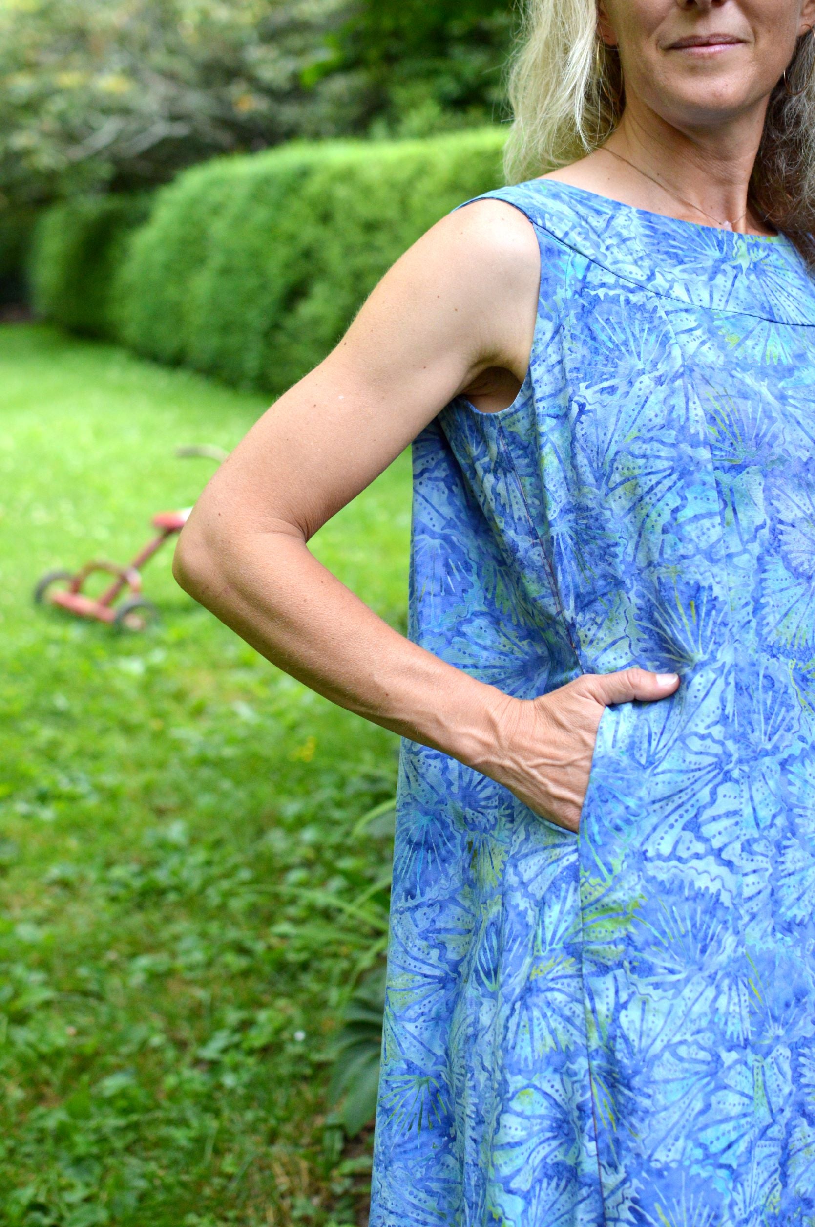 White woman wearing a sleeveless blue batiked muumuu, standing outside by shrubbery.  her hand is in her pocket and view is close up on the pocket.