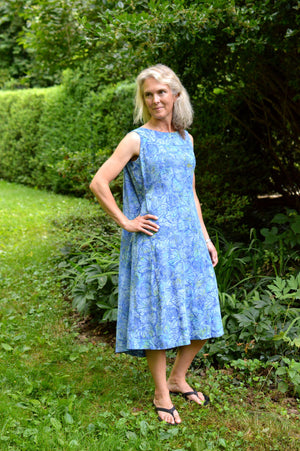 White woman wearing a sleeveless blue batiked muumuu, standing outside by shrubery.