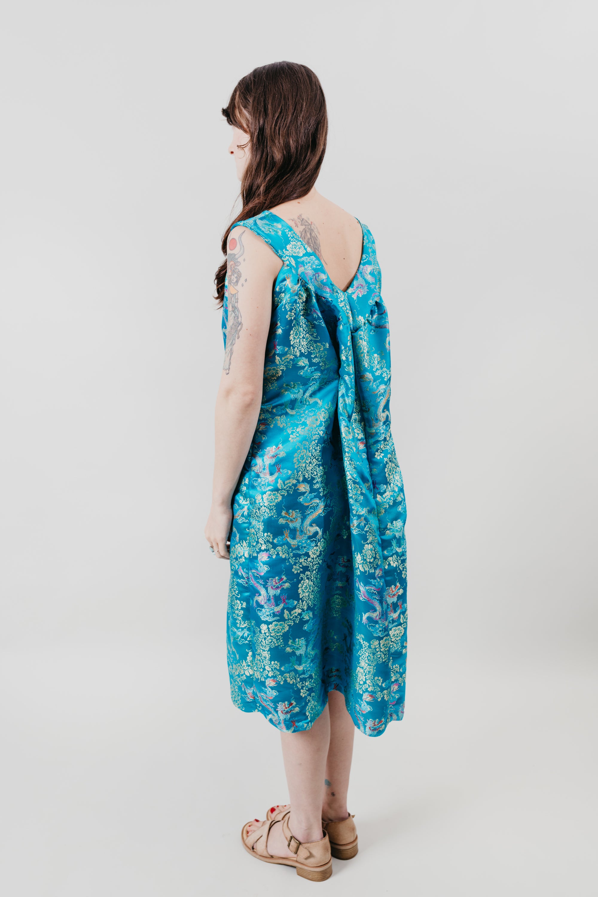 Woman standing with her back to the camera in front of a white backdrop in a sleeveless teal brocade muumuu.