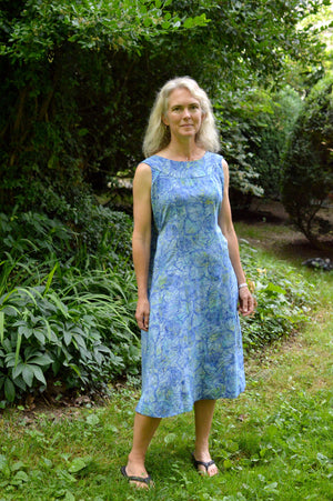 White woman wearing a sleeveless blue batiked muumuu, standing outside by shrubery.