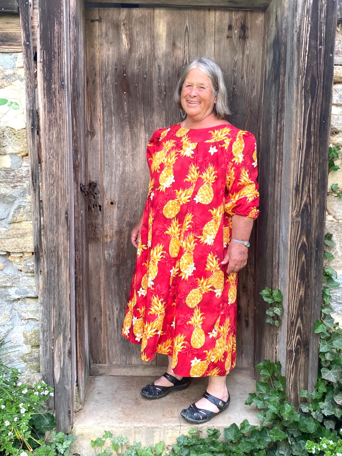Older woman wearing a red muumuu with yellow pineapples on it.  Sleeves are 3/4 length and she is standing in a wooden doorway.