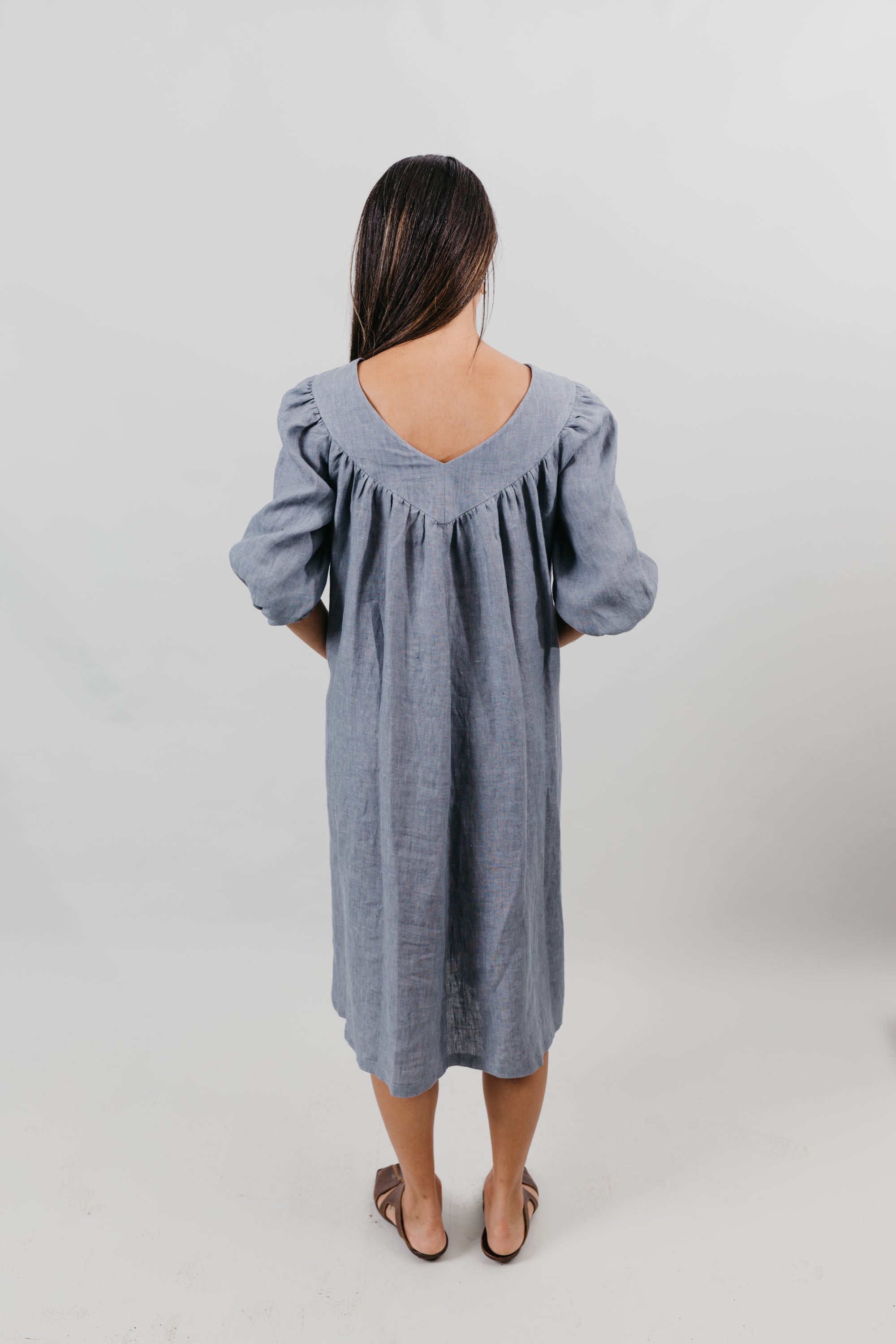Back view of woman wearing a grey linen muumuu with 3/4 length sleeves.  Background is white backdrop.