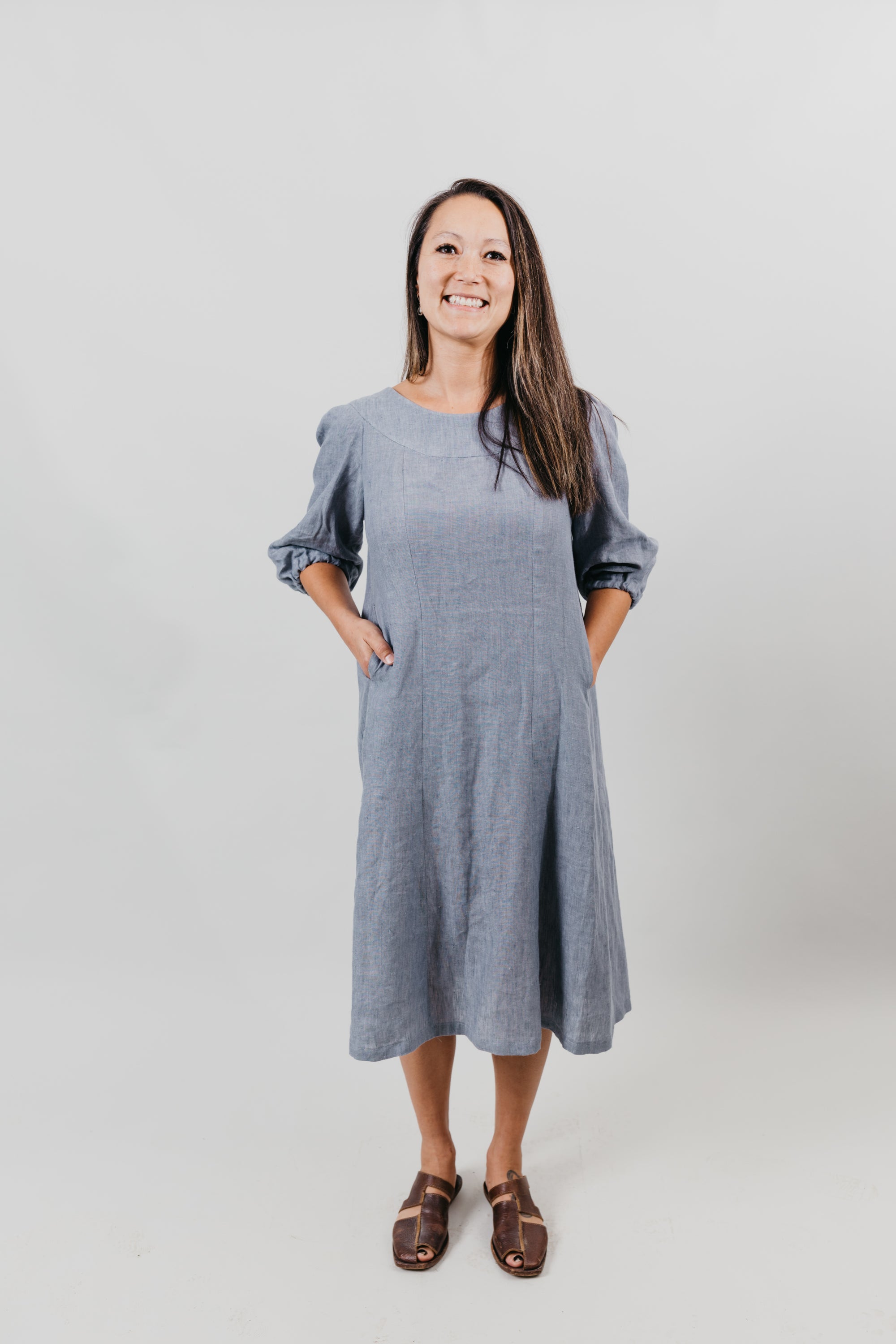 Grey linen muumuu on an Asian American woman standing in front of a white backdrop.