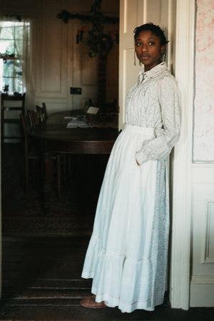 Photo of young woman standing in a doorway wearing prairie dress and apron.  model rests back on door frame and has her hands in the pockets of the apron.