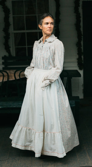 Photo of woman standing outdoors wearing prairie dress.  Model is truning and skirt is full.  Hands are in the pockets of the apron.