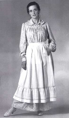 Blacka nd white photo of model wearing prairie dress and apron with studio backdrop.