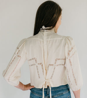 Back view of brunette woman standing in front of a white studio backdrop, wearing 205 Gibson Girl Blouse with hand left hand on her hip.