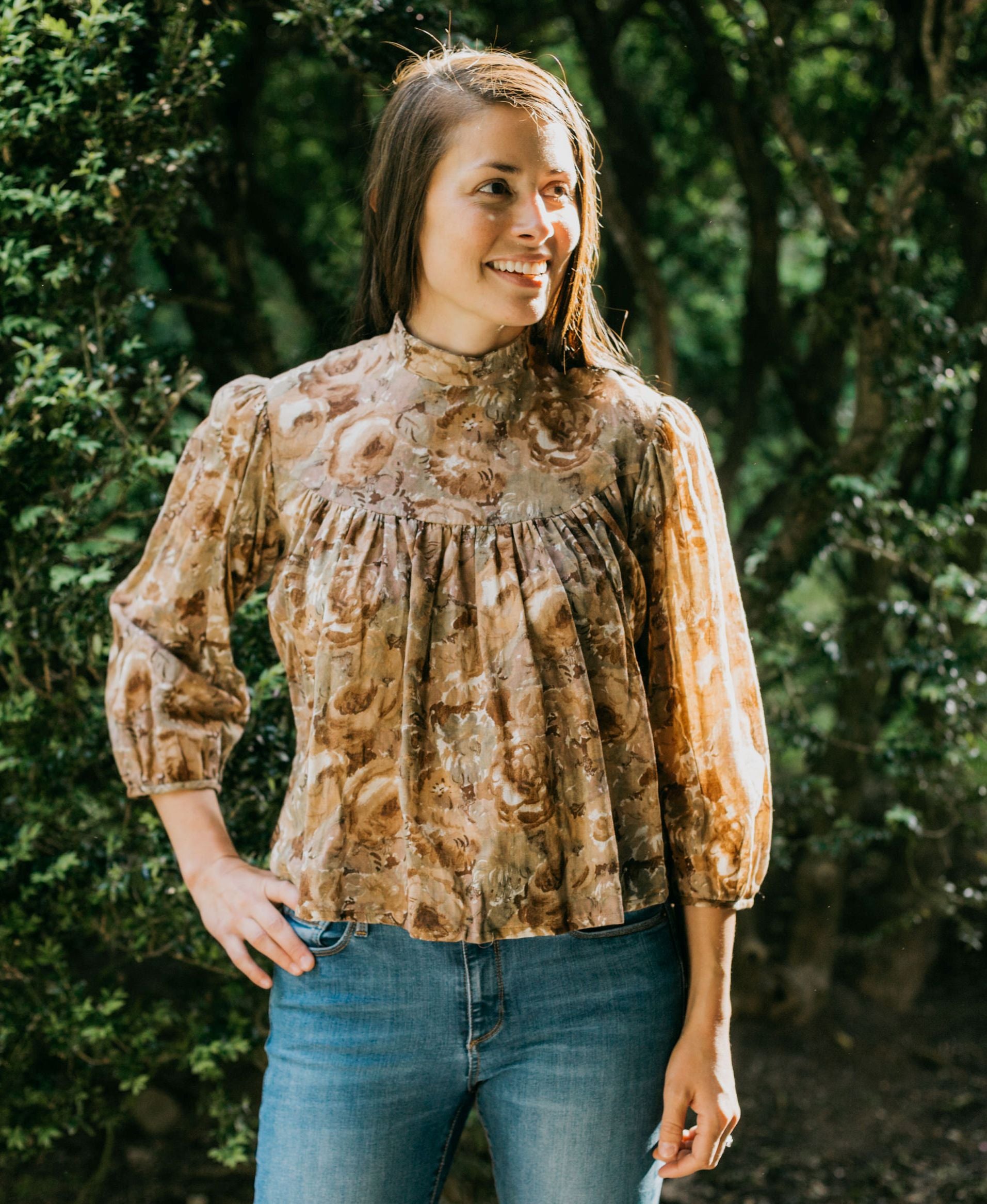 Brunette woman standing surrounded by greenery with hand on her hip looking to the right, wearing 205 Gibson Girl Blouse.