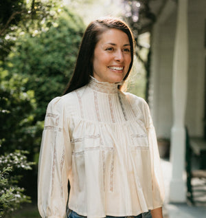 Brunette woman smiling looking to the right,  surrounded by greenery wearing 205 Gibson Girl Blouse.