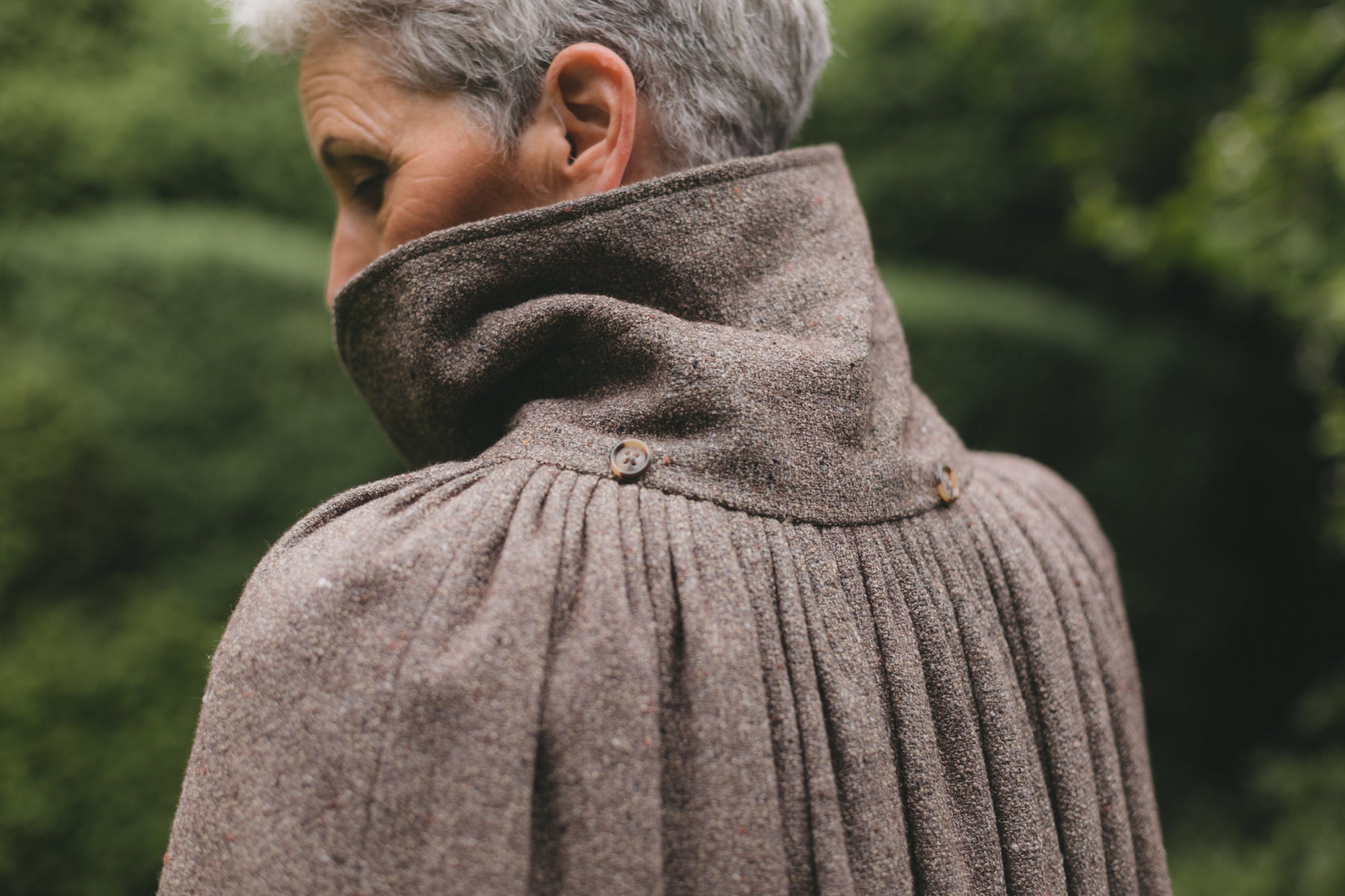 Back close up view of older white woman with gray hair surrounded by greenery wearing 207 Kinsale Cloak with the collar upturned.