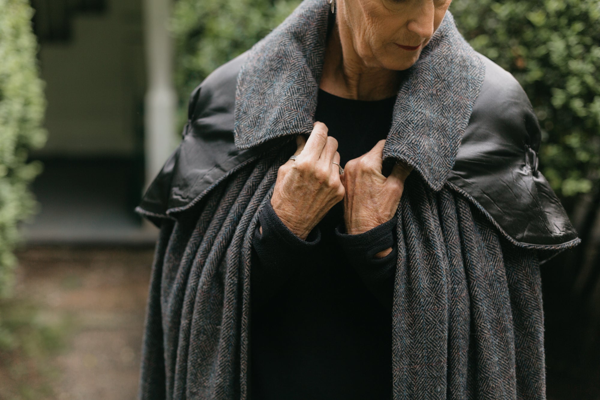 Close up of older white woman standing surrounded by greenery wearing 207 Kinsale Cloak holding the each side of the collar with hands close to chest.