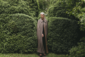 Older white woman with gray hair standing surrounded by greenery wearing 207 Kinsale Cloak without a hood.