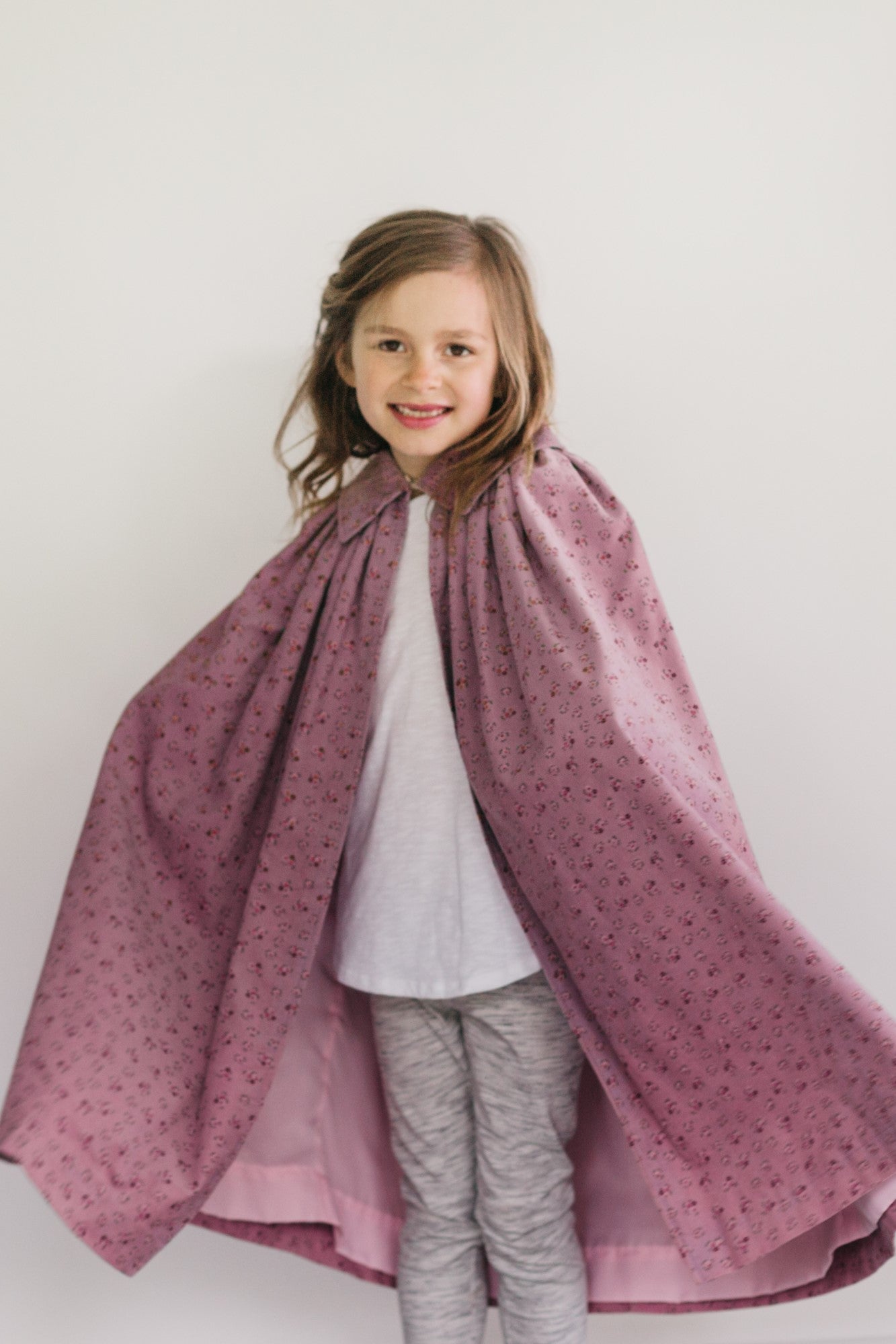 Smiling small girl standing in front of a white studio backdrop wearing 208 Kinsale Cloak for Young Maidens arms extended to either side to display the voluminous cloak.