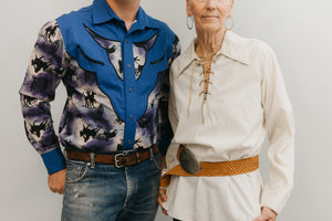 A man and a older woman standing in front of a white studio backdrop ,wearing 212 Five Frontier Shirts. Man wearing View D with decorative bull's head yoke and woman wearing View A with lace up front with belt around the waist. 