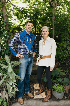Man and an older woman standing in a pathway surrounded by greenery both wearing 212 Five Frontier Shirts. Man is wearing  View D with decorative bull's head yoke, woman is wearing View A with lace up front and a belt around the waist.