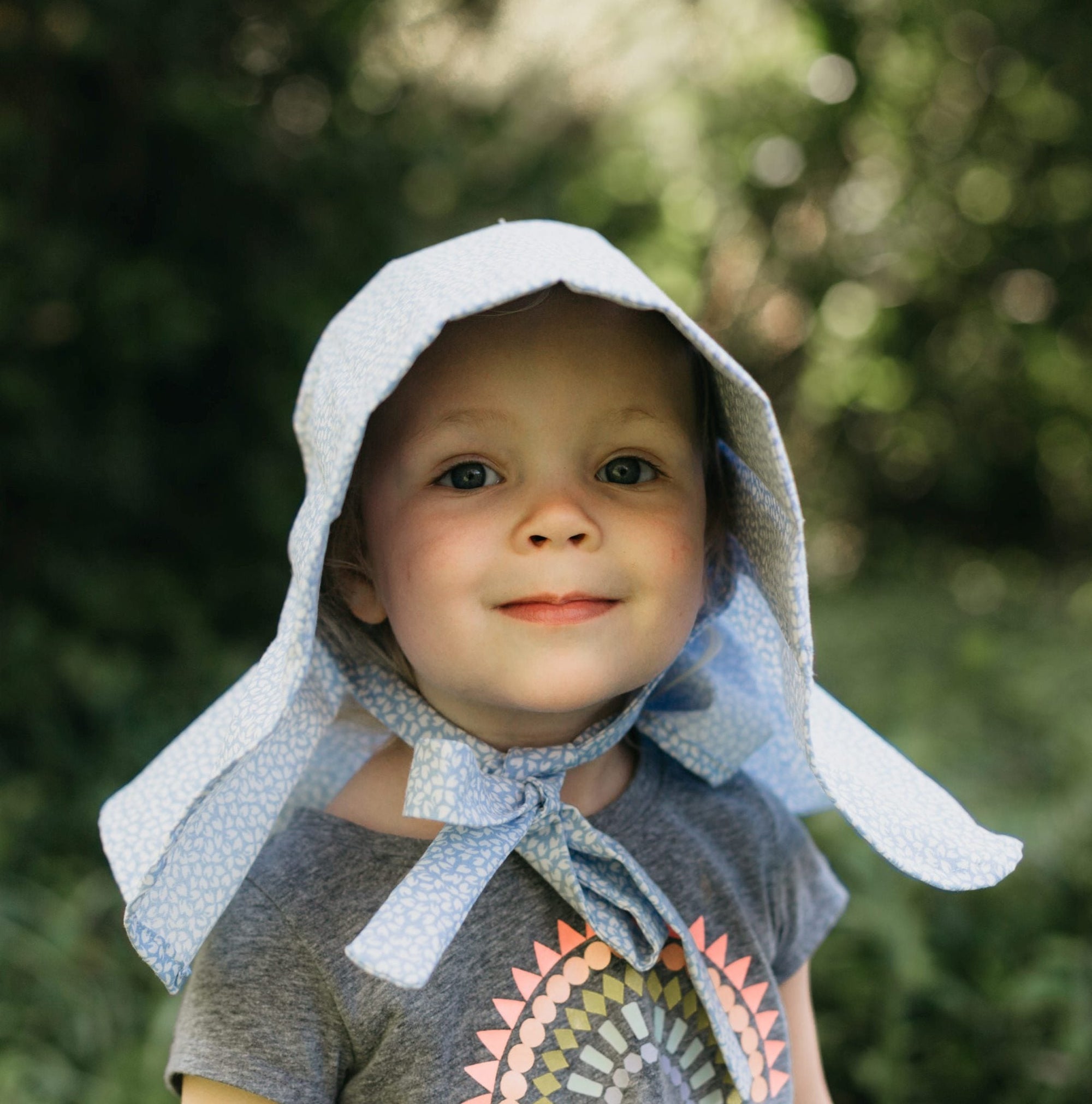 Little girl surrounded by greenery wearing 213 Sunbonnet.