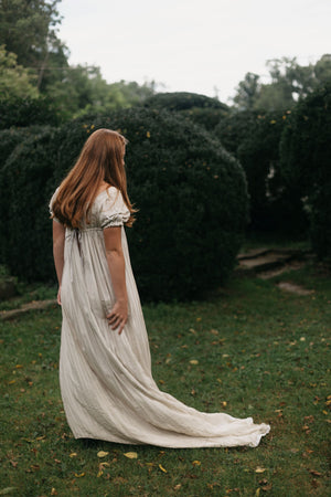 Back view of red head young woman standing surrounded by greenery wearing 215 Empire Dress.