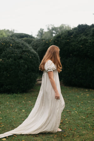 Side view of red head young woman standing surrounded greenery wearing 215 Empire Dress. 