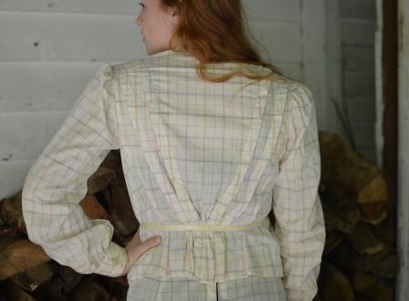 Back view of young red head woman standing with one hand on her hip wearing 216 Schoolmistresses Shirtwaist and skirt. the shirt is gathered in the back and tied at the waist.